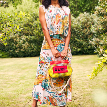Woman in a floral dress carrying a colorful CLRS by SAAT bag while standing in a garden.